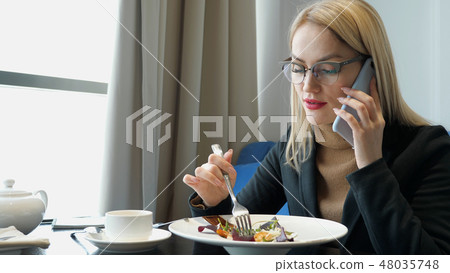 Young blonde woman manager talking on the phone and eating at the cafe. 48035748