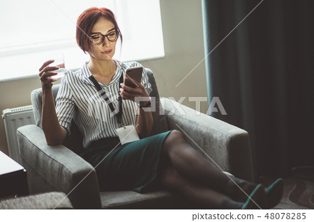 Businesswoman sits in armchair with water glass in hand and talking on phone 48078285