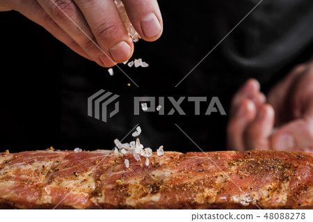 Raw piece of meat, beef ribs. The hand of a male chef puts salt and spices on a dark background. 48088278