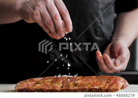 Raw piece of meat, beef ribs. The hand of a male chef puts salt and spices on a dark background. 48088279