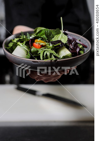 Cook man in a black apron holds a dish of salad in his hand. Against the background of the kitchen 48088654