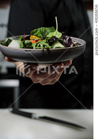 Cook man in a black apron holds a dish of salad in his hand. Against the background of the kitchen Cook man in a black apron holds a dish of salad in his hand. Against the background of the kitchen 48088656