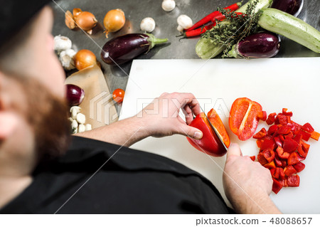 Male chef cuts vegetables for salad in a restaurant in a black apron. White cutting board, closeup 48088657