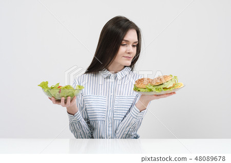 Slender brunette girl on a white background chooses between a plate of fast food and hamburgers and Slender brunette girl on a white background chooses between a plate of fast food and hamburgers and 48089678