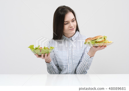 Slender brunette girl on a white background chooses between a plate of fast food and hamburgers and Slender brunette girl on a white background chooses between a plate of fast food and hamburgers and 48089681