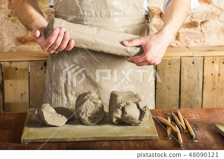 the man wearing apron and shirt holding a big piece of clay, on the work table lying board, pieces 48090121