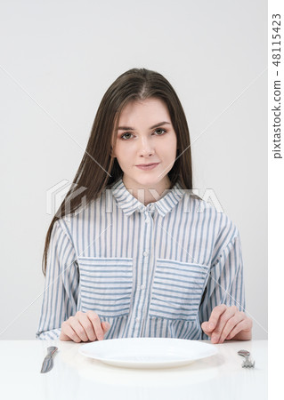 Hungry thin girl sitting at the table in front of an empty plate with a knife and fork. 48115423