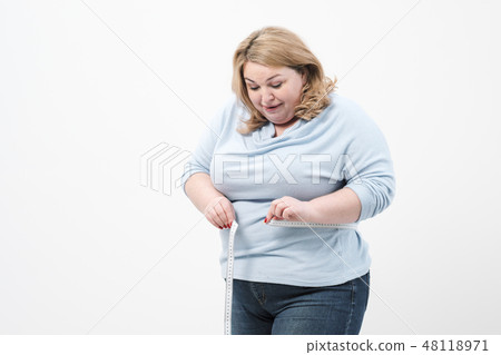 A fat woman measures her waist with a measuring tape in casual clothing on a white background. A fat woman measures her waist with a measuring tape in casual clothing on a white background. 48118971