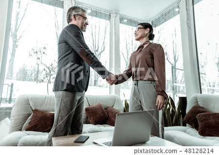 Dark-haired elegant woman in grey trousers and a man in eyeglasses shaking hands Dark-haired elegant woman in grey trousers and a man in eyeglasses shaking hands 48128214