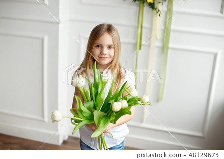 Portrait of a very cute pretty blonde girl in a white shirt against a white wall with a bouquet of 48129673