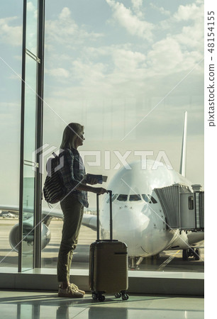 Woman with boarding passes and hand baggage looks out the terminal window on a large airliner 48154155