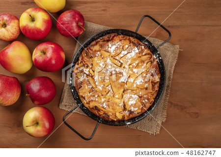 A photo of an apple pie in a pan, shot from the top on a dark rustic wooden background with apples A photo of an apple pie in a pan, shot from the top on a dark rustic wooden background with apples 48162747