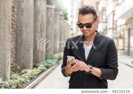 Outdoors leisure. Young man in suit and sunglasses standing on city street using app on smartphone Outdoors leisure. Young man in suit and sunglasses standing on city street using app on smartphone 48238139