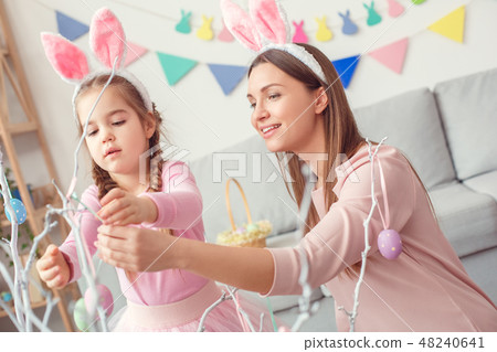 Mother and daughter in bunny ears together at home easter concept hanging eggs on tree Mother and daughter in bunny ears together at home easter concept hanging eggs on tree 48240641
