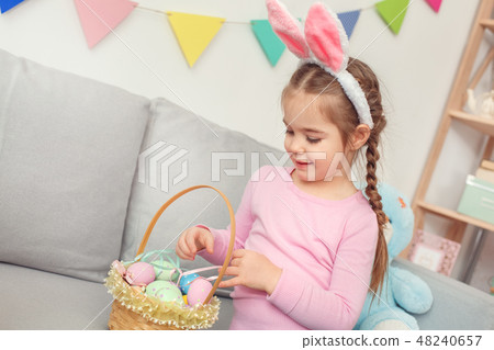 Little cute girl in bunny ears at home easter celebration concept looking at basket with eggs Little cute girl in bunny ears at home easter celebration concept looking at basket with eggs 48240657