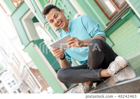 Outdoors leisure. Young man sitting on city street playing game on digital tablet smiling excited Outdoors leisure. Young man sitting on city street playing game on digital tablet smiling excited 48242463