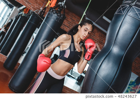 Young woman in boxing gloves in gym standing breathing before kicking punching bag 48243627