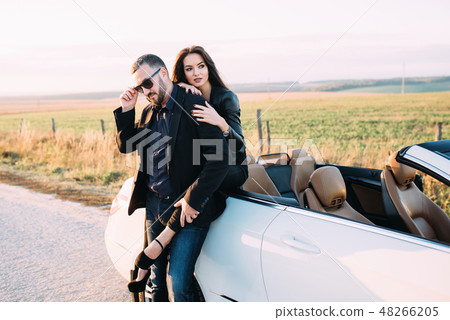 Sympathetic couple husband and wife spend time outside the city, sitting on a car hood Sympathetic couple husband and wife spend time outside the city, sitting on a car hood 48266205
