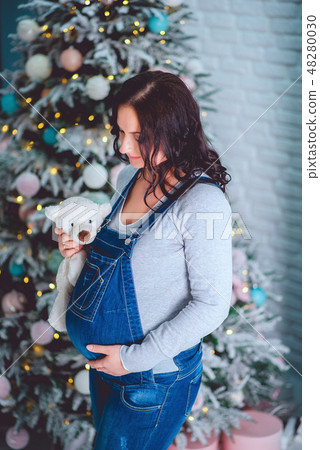 Beautiful young pregnant girl in blue denim overalls holding a toy 48280030