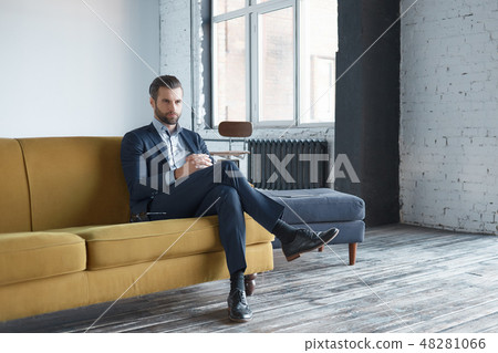 Business look: successful and handsome business man is sitting on office sofa and looking aside Business look: successful and handsome business man is sitting on office sofa and looking aside 48281066