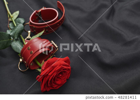 Erotic toys. Top view of red leather handcuffs and rose against of black silk fabric. 48281411