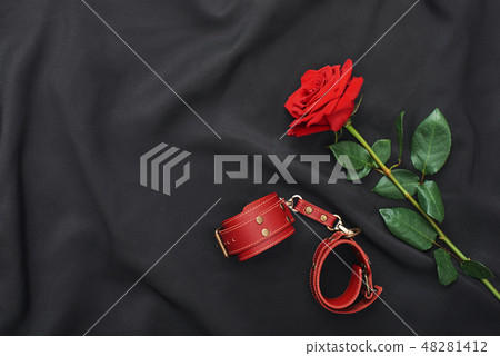 BDSM fantasy play. Top view of red leather handcuffs and rose against of black silk fabric 48281412