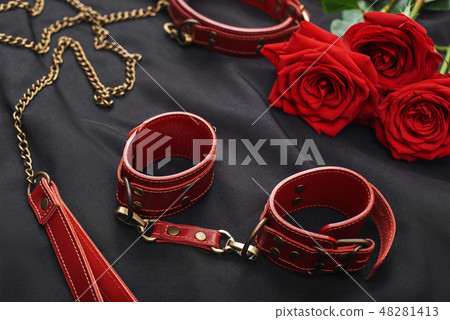 Erotic games. Top view of BDSM leather stuff: red handcuffs and roses against of black silk fabric 48281413