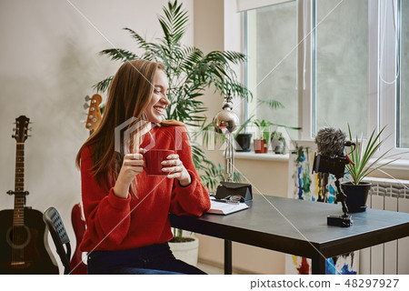 Successful blogger. Side view of smiling female blogger drinking a tea while making a new video at Successful blogger. Side view of smiling female blogger drinking a tea while making a new video at 48297927