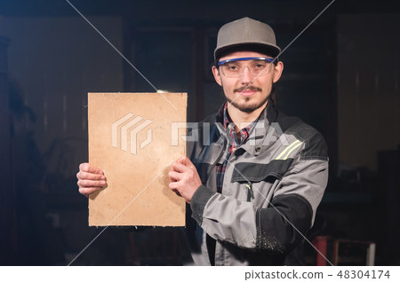Portrait of a young carpenter in overalls and goggles with a mock up board holding a blank sign in 48304174