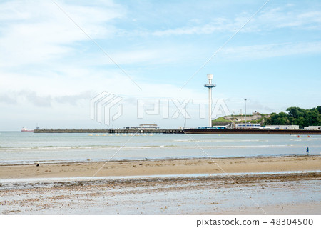 A shallow sand under grassy blue sky with white clouds and a spacious beach of gravel and a street tower and a tower that can be seen far away 48304500
