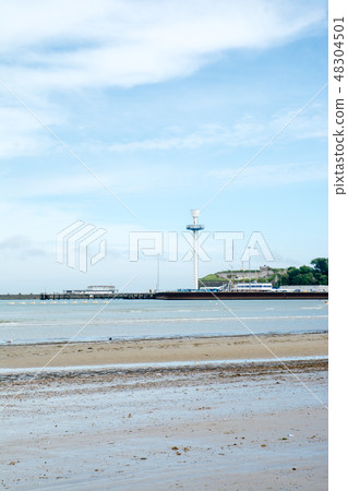A shallow sand under grassy blue sky with white clouds and a spacious beach of gravel and a street tower and a tower that can be seen far away 48304501