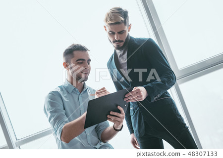 Two focused male co-workers deep in discussion together, making notes, smiling, while standing in a Two focused male co-workers deep in discussion together, making notes, smiling, while standing in a 48307133
