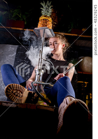 Young, beautiful woman smokes a hookah with pineapple in cafe. shod in rough men's shoes. It 48329245
