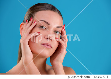 Close up Beauty portrait of young woman brunette smiling and touching her face on blue background Close up Beauty portrait of young woman brunette smiling and touching her face on blue background 48337801