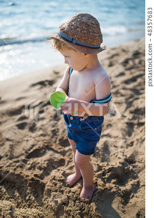 A small toddler boy standing on beach on summer holiday, playing. A small toddler boy standing on beach on summer holiday, playing. 48357683