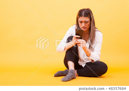 Girl looking worried at her phone while sitting down over yellow background 48357684