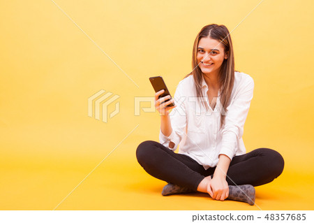 Beautiful girl smiling her phone while sitting down over yellow background Beautiful girl smiling her phone while sitting down over yellow background 48357685