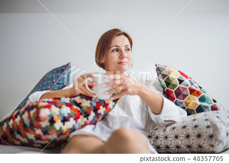 Young woman with night shirt sitting indoors on bed in the morning, holding cup. Young woman with night shirt sitting indoors on bed in the morning, holding cup. 48357705