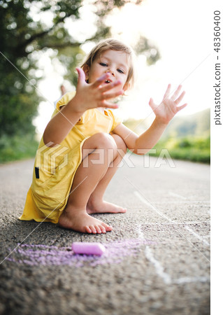 Small cute girl on a road in countryside in sunny summer nature, drawing with chalk. 48360400