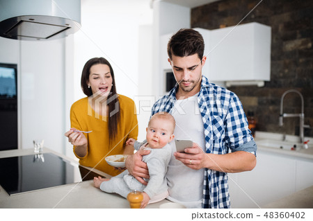 A young family at home, a man holding a baby and a woman feeding her. 48360402
