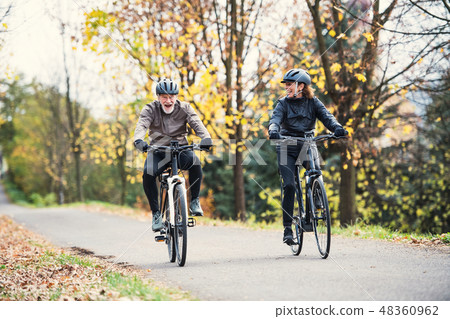 A senior couple with electrobikes cycling outdoors on a road in park in autumn. A senior couple with electrobikes cycling outdoors on a road in park in autumn. 48360962