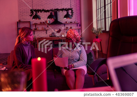African american plump fortune-teller in ethnic headwear sitting on the sofa with the client 48369807
