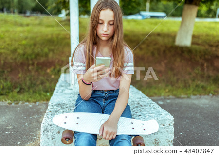 Teen girl 12-13 years old, sitting in hands of a skate. In summer in city in casual jeans and pink T Teen girl 12-13 years old, sitting in hands of a skate. In summer in city in casual jeans and pink T 48407847
