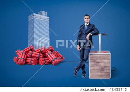 Businessman standing next to detonator box connected to heap of dynamite beside high-rise building Businessman standing next to detonator box connected to heap of dynamite beside high-rise building 48433542