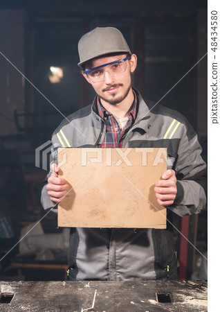 Portrait of a young carpenter in overalls and goggles with a mock up board holding a blank sign in 48434580