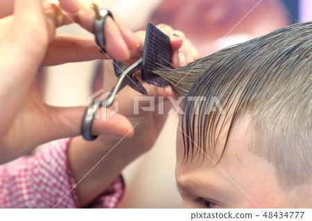 Hairdresser cuts bangs with scissors on boy's head. Stylist's hands close-up, side view. Hairdresser cuts bangs with scissors on boy's head. Stylist's hands close-up, side view. 48434777