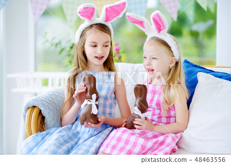 Two cute little sisters wearing bunny ears eating chocolate Easter rabbits 48463556
