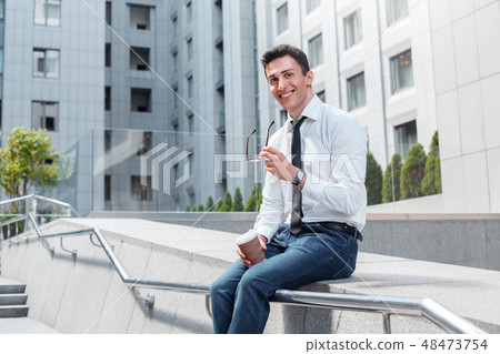 Business lifestyle. Businessman with eyeglasses sitting on fence on the city street holding cup of 48473754