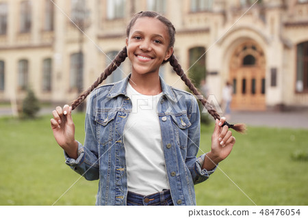 Young schoolgirl in jeans standing at school yard holding braids looking camera playful Young schoolgirl in jeans standing at school yard holding braids looking camera playful 48476054