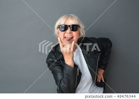 Senior woman in leather jacket and sunglasses studio standing isolated on gray showing tongue Senior woman in leather jacket and sunglasses studio standing isolated on gray showing tongue 48476344